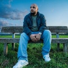 Ein Mann sitzt auf einer Bank im Freien, umgeben von Gras und einem bewölkten Himmel.