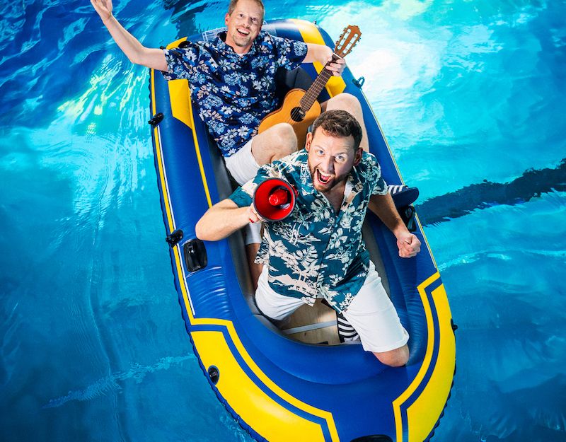Zwei Männer in Hawaiihemden in einem Schlauchboot im Pool, einer mit Gitarre, der andere mit Megafon.