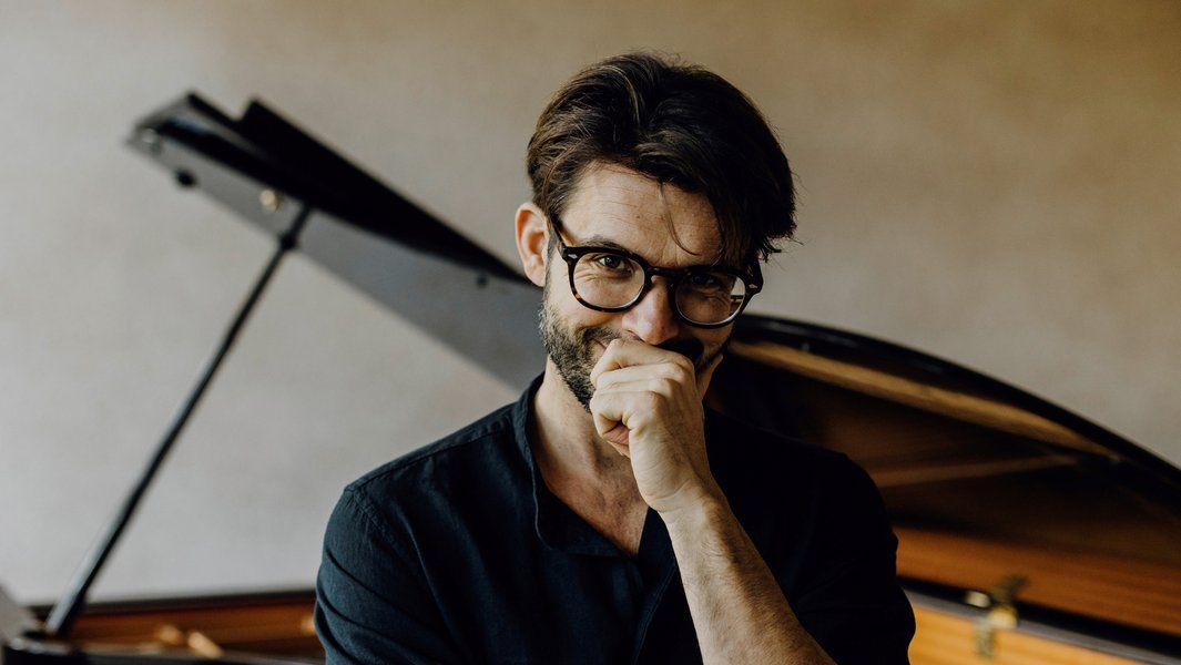 Ein Mann mit Brille steht vor einem Flügel und lächelt nachdenklich.
