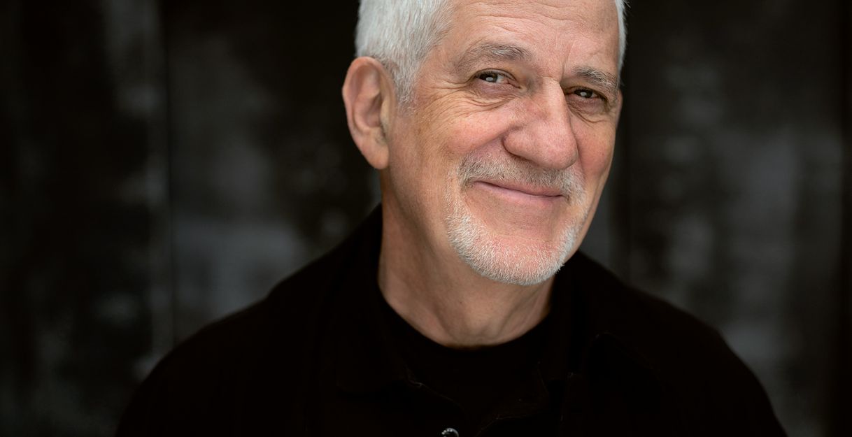 Ein älterer Mann mit weißem Haar und Bart lächelt in die Kamera vor einem dunklen Hintergrund.