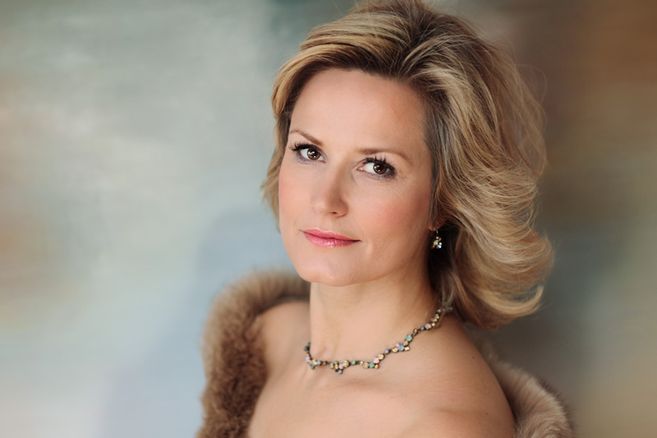 Porträt einer Frau mit blondem Haar und elegantem Schmuck vor einem unscharfen Hintergrund.