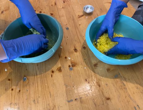 Zwei Personen mit blauen Handschuhen mischen bunte Zutaten in Schüsseln auf einem Holztisch.