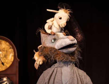 Eine Puppenfigur eines Wolfes mit einem kleinen Ziegenbock auf dem Kopf, vor einem dunklen Hintergrund und einer Uhr.