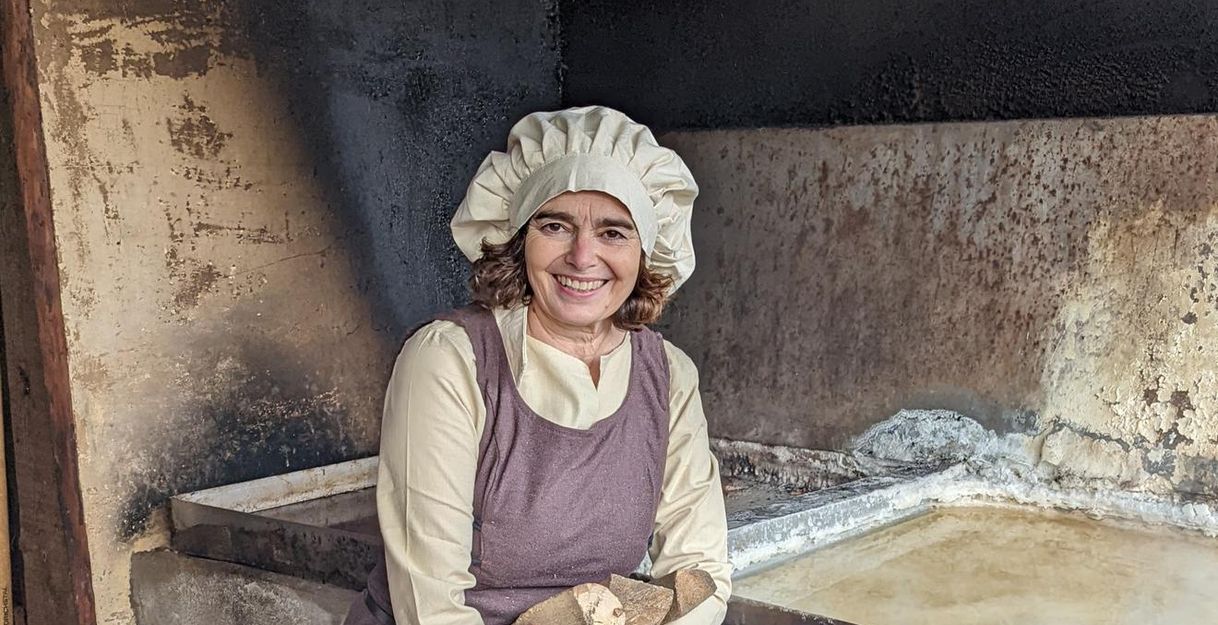 Frau in historischer Kleidung mit Holzscheiten vor einem alten Salzsiedebecken.