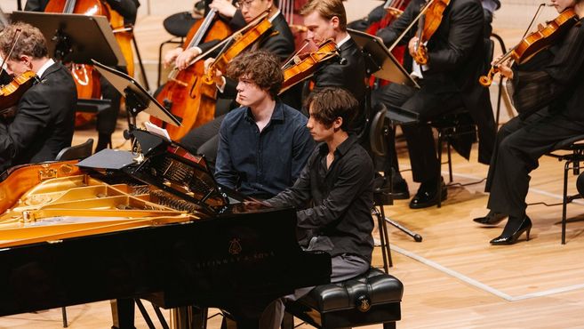 Zwei Pianisten spielen auf einem Flügel, umgeben von einem Orchester mit Streichinstrumenten.