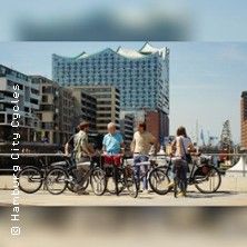 Radfahrer stehen auf einer Brücke in Hamburg mit Blick auf die Elbphilharmonie und die Speicherstadt; im Hintergrund sind moderne Gebäude und Hafenkräne zu sehen.