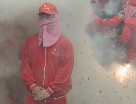 Sie sehen ein Filmstill aus der Arbeit HEAT von Yalda Afsah. Im Mittelpunkt ist das Porträt eines Teilnehmers der taiwanischen Feuerwerkszeremonie zu sehen. Die Person trägt eine Brille und hat ein Tuch vor Mund und Nase. Dichter Rauch umgibt ihren Kopf und Oberkörper.