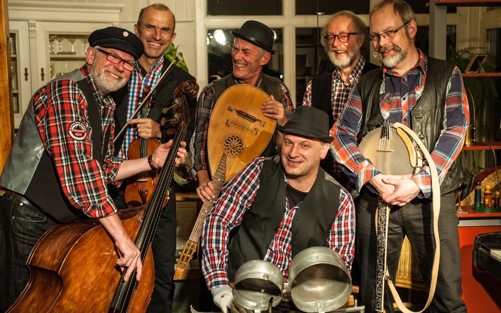 Eine Gruppe von sechs Männern in karierten Hemden und Westen posiert mit Musikinstrumenten in einem Raum.