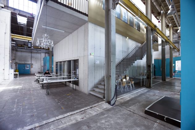 Innenansicht einer industriellen Halle mit Tischen, Stühlen und Treppe.