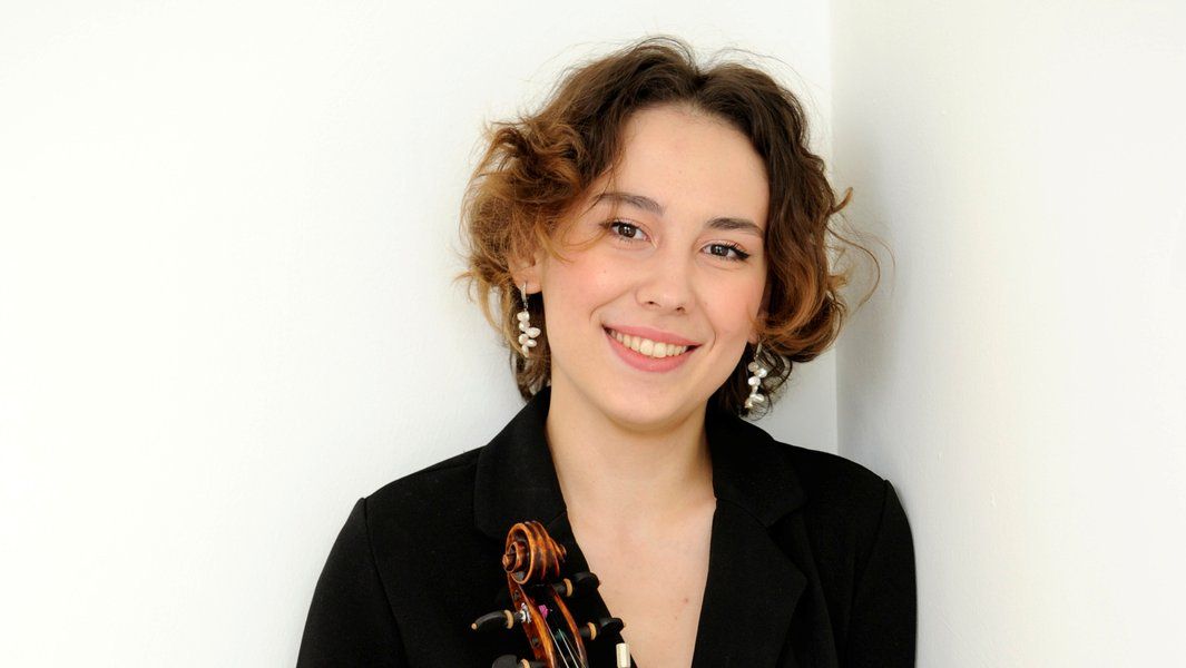 Eine lächelnde Frau mit kurzen, lockigen Haaren hält eine Geige. Sie trägt einen schwarzen Blazer und auffällige Ohrringe.