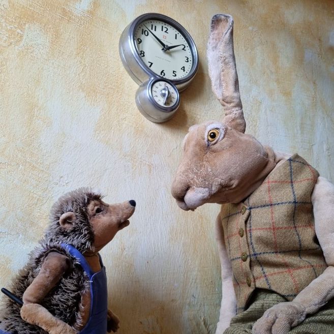 Ein Plüschhase und ein Plüschigel stehen vor einer Wanduhr und schauen sich an.