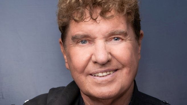 Porträt eines lächelnden Mannes mit lockigem Haar vor einem grauen Hintergrund.