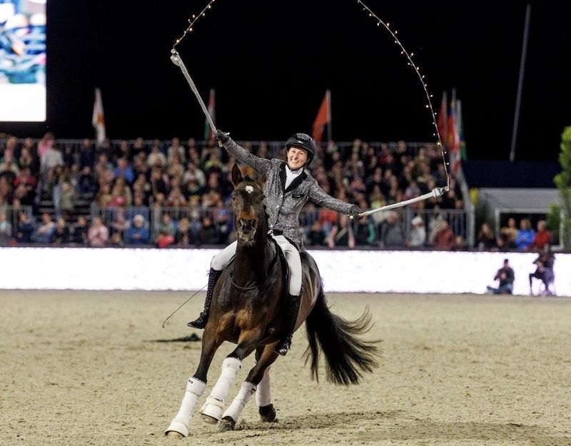 Reiterin auf einem Pferd bei einer Show, schwingt eine lange Peitsche, umgeben von einem Publikum.