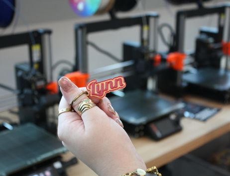 Hand hält einen kleinen 3D-gedruckten Anhänger mit der Aufschrift „Lynn“ vor mehreren 3D-Druckern.
