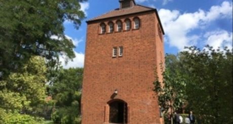 Ein roter Backsteinturm mit mehreren Fenstern vor einem blauen Himmel mit weißen Wolken.