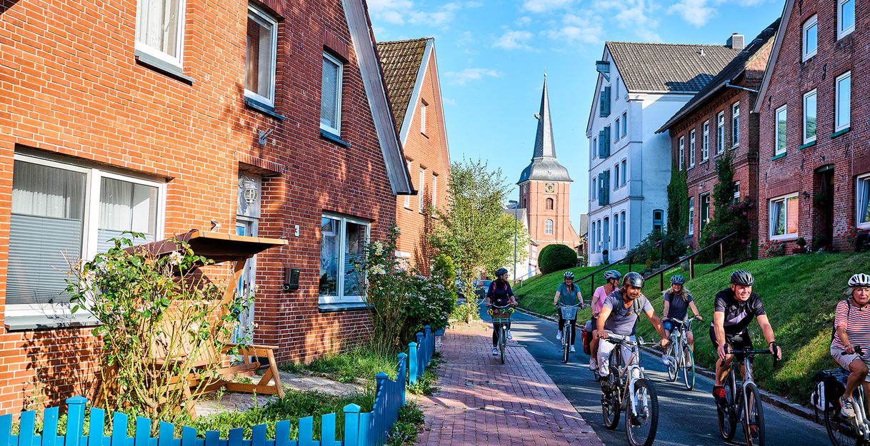 Gruppe von Radfahrern auf einer Straße mit Backsteinhäusern und Kirchturm im Hintergrund.
