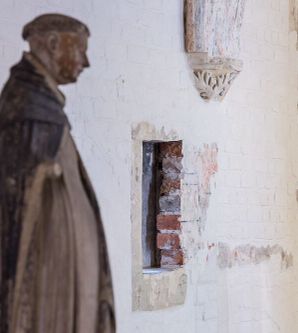 Foto in schräger Perspektive auf eine weiß gestrichene Backsteinwand., in die ein kleiner Durchbruch geschlagen wurde. Im Vordergrund steht eine Dominikanerstatue.