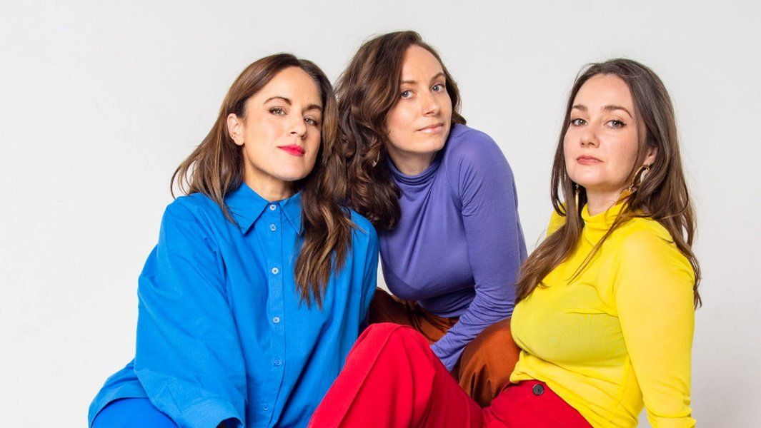 Drei Frauen in bunter Kleidung posieren vor einem weißen Hintergrund.