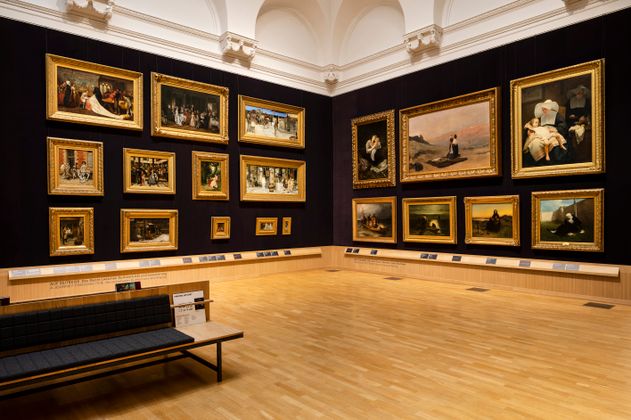 Innenansicht eines Museumsraums mit Gemälden an der Wand und einer Bank im Vordergrund.