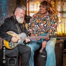 Ein Mann mit Gitarre und eine Frau sitzen zusammen und lachen in einem gemütlichen Raum.