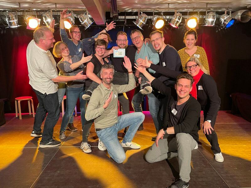 Eine Gruppe von Menschen posiert fröhlich auf einer Bühne mit bunten Lichtern im Hintergrund.