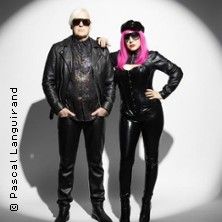 Zwei Personen in schwarzer Lederkleidung und Sonnenbrillen posieren vor einem weißen Hintergrund.