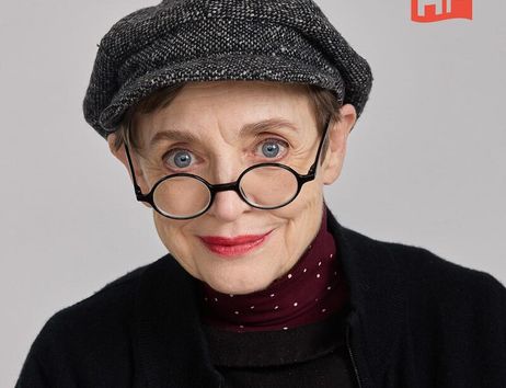 Eine ältere Frau mit Brille und Mütze lächelt in die Kamera. Sie trägt roten Lippenstift und einen schwarzen Pullover.