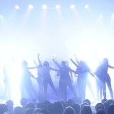 Silhouetten von Menschen auf einer Bühne mit hellem Licht im Hintergrund.
