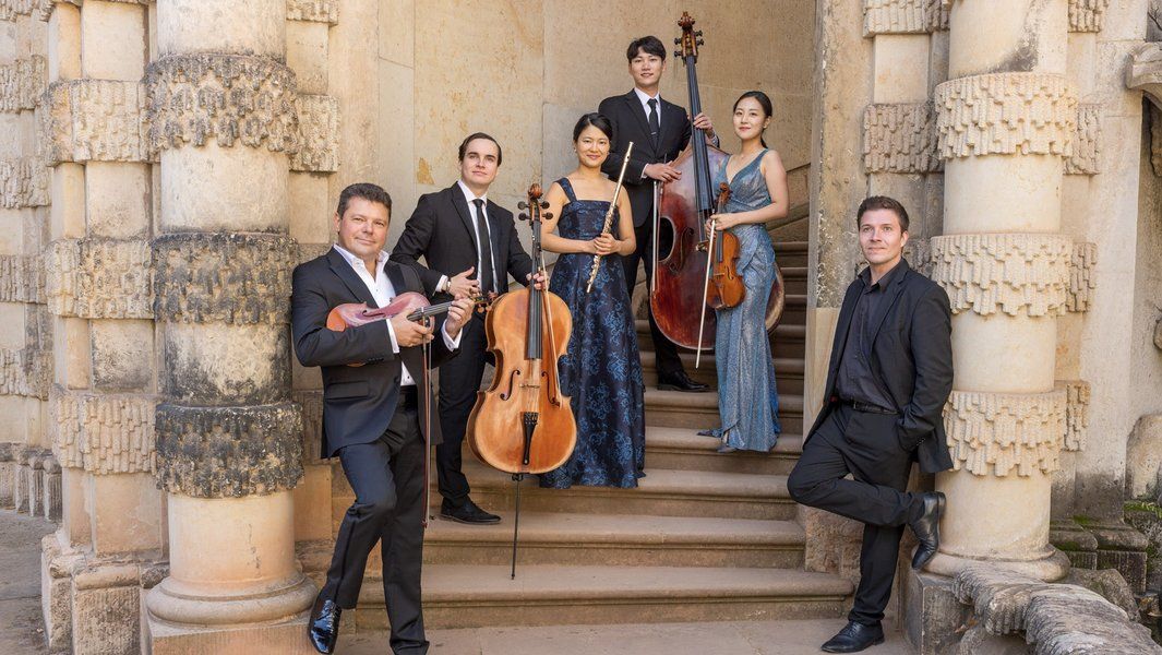 Eine Gruppe von sechs Musikern in eleganter Kleidung posiert mit ihren Instrumenten vor einer historischen Kulisse.