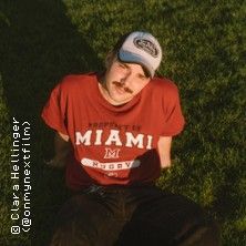 Person mit rotem Miami-Shirt und Baseballkappe sitzt auf Gras.