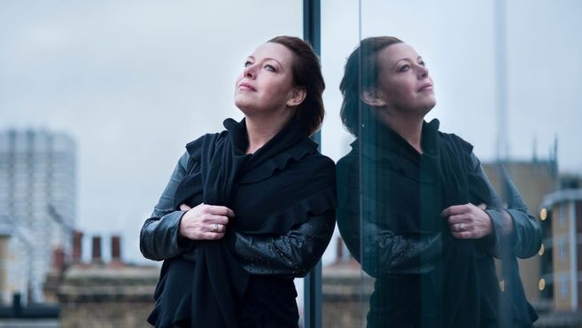 Eine Frau lehnt an einer Glaswand und schaut nachdenklich in die Ferne. Ihr Spiegelbild ist im Glas zu sehen.