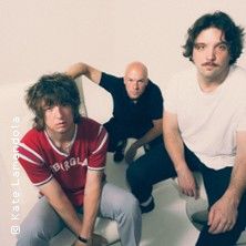 Drei Männer posieren in einem hellen Raum, einer sitzt auf einem Sofa, die anderen beiden stehen daneben.