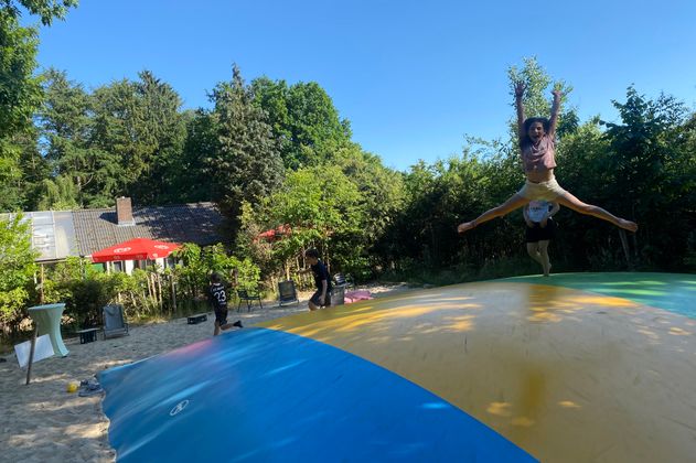 Kinder springen auf einem großen Hüpfkissen. Das Hüpfkissen ist gestreift und umgeben von Sand und Bäumen. Die Sonne schient, der Himmel ist blau