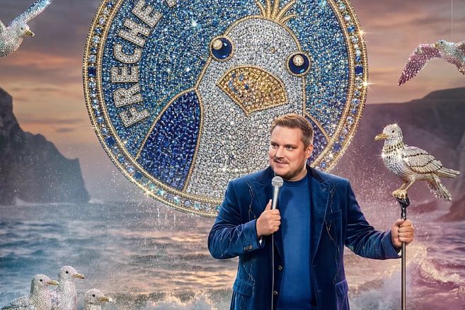 Ein Mann hält ein Mikrofon und steht vor einem großen, glitzernden Möwen-Logo. Im Hintergrund sind das Meer und Klippen zu sehen.