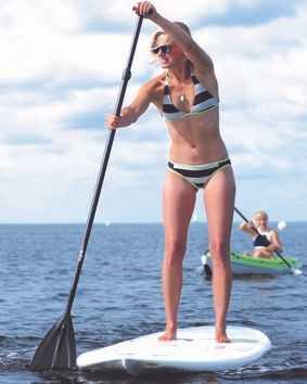 Eine Frau steht auf einem Stand-Up-Paddleboard auf dem Wasser, während eine andere Person in einem Kajak im Hintergrund paddelt.