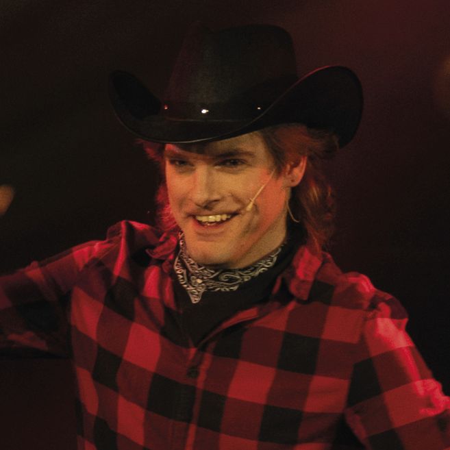 Person mit Cowboyhut und kariertem Hemd auf einer Bühne.