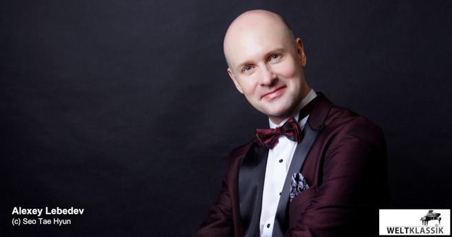 Ein Mann in einem eleganten Anzug mit Fliege lächelt in die Kamera vor einem dunklen Hintergrund.