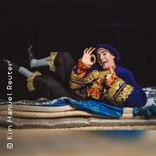 Ein Schauspieler in einem bunten Kostüm liegt auf mehreren Matratzen und posiert fröhlich.