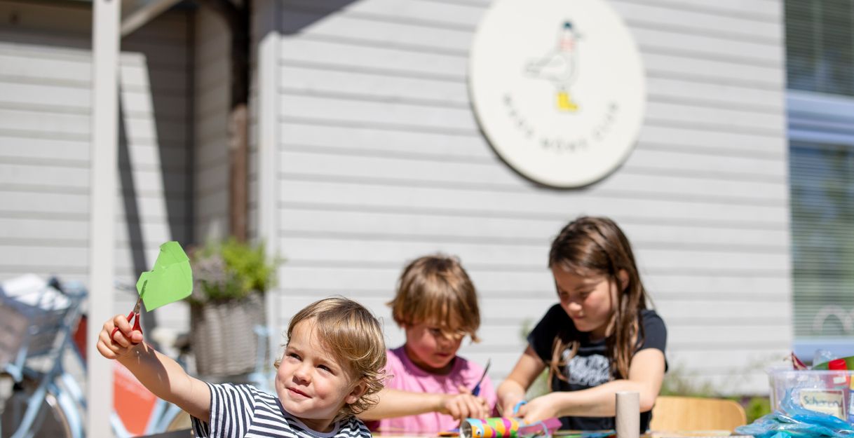 Kinder basteln an einem Tisch im Freien vor einem Gebäude mit einem Schild des Mads Möwe Clubs.