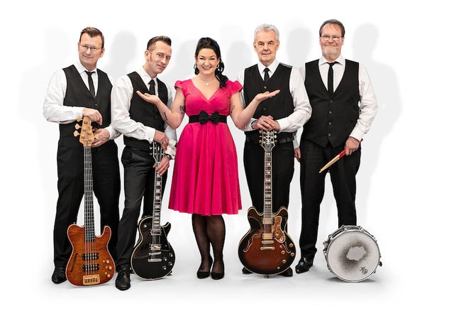 Eine Band mit vier Männern und einer Frau in einem pinken Kleid posiert mit Instrumenten.
