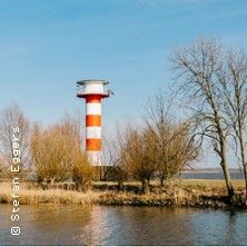 Ein rot-weiß gestreifter Leuchtturm steht hinter Bäumen am Ufer eines Gewässers.