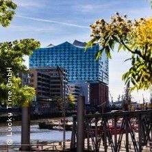 Blick auf die Elbphilharmonie in Hamburg, umgeben von Bäumen und einem Fluss im Vordergrund.