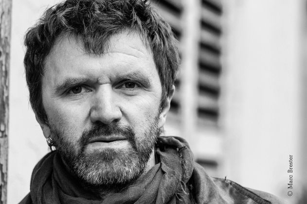 Schwarz-weißes-Portraitfoto einer männlich gelesenen Person mittleren Alters mit Bart. Sie trägt einen Schal, mehrere Ohrringe im einen Ohr und schaut ernst.