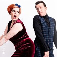 Zwei Personen in auffälliger Pose, eine Frau in rotem Samtkleid und ein Mann in kariertem Anzug, vor weißem Hintergrund.