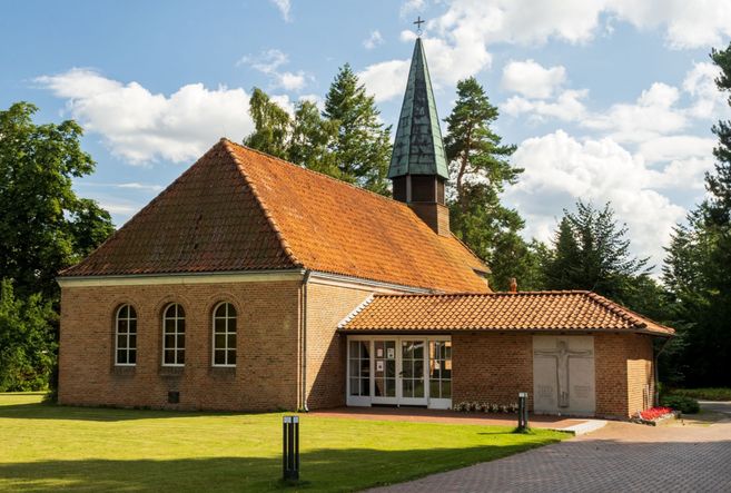 Kleine Backsteinkirche mit rotem Ziegeldach und schlankem Turmkreuz, umgeben von Bäumen und Rasen.
