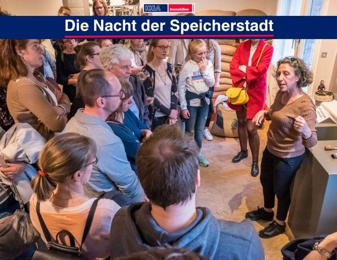 Eine Gruppe von Menschen hört einer Frau in einem Raum mit Kaffeesäcken zu. Oben steht 'Die Nacht der Speicherstadt'.
