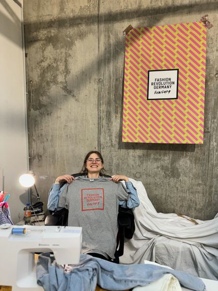 Foto einer Nähwerkstatt mit Nähmaschine im Vordergrund und junger Frau, die lächelnd ein graues T-Shirt hochhält auf dem "Fashion Revolution Germany" steht.  