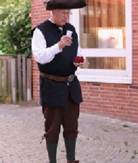 Ein Mann in historischer Kleidung steht auf einem gepflasterten Weg vor einem Backsteingebäude.