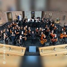Ein Orchester sitzt in einem Halbkreis in einer Kirche, bereit für ein Konzert. Die Musiker halten ihre Instrumente und blicken zur Kamera.