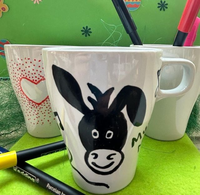 Bemalte Tassen mit Eselmotiv und Herzen, umgeben von Porzellanmalstiften auf grünem Hintergrund.